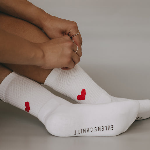 Frau trägt weiße Socken mit rotem Herzmotiv auf den Fersen – Nahaufnahme der Beine und Socken mit Händen und filigranem Schmuck.