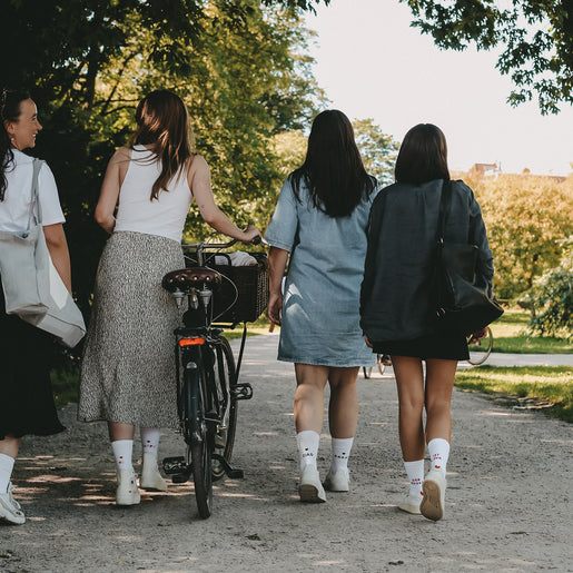Rückansicht von vier Frauen mit weißen Socken mit Schriftzügen wie ‚CIAO KAKAO‘ und ‚DER BODEN IST LAVA‘ beim Spaziergang im Park.