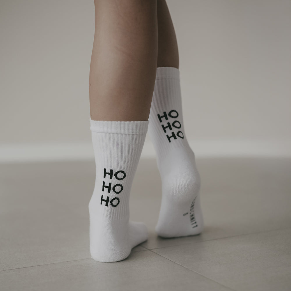 Nahaufnahme von Beinen mit weißen Socken mit schwarzem ‚HO HO HO‘ Schriftzug. Die Person steht auf hellem Boden.