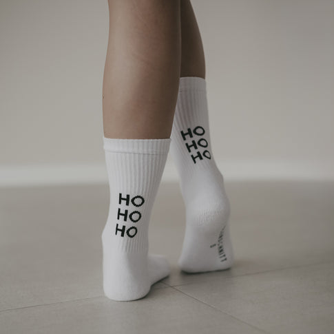Nahaufnahme von Beinen mit weißen Socken mit schwarzem ‚HO HO HO‘ Schriftzug. Die Person steht auf hellem Boden.
