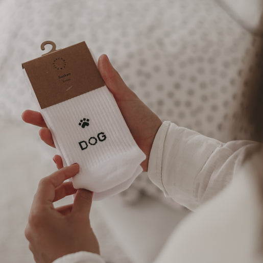 Eine Person in weißem Oberteil hält ein Paar weiße Socken mit einem schwarzen Pfotenabdruck und dem Wort „DOG“ in der Hand; die Socken sind mit einer braunen Kartonaufhängung verpackt.