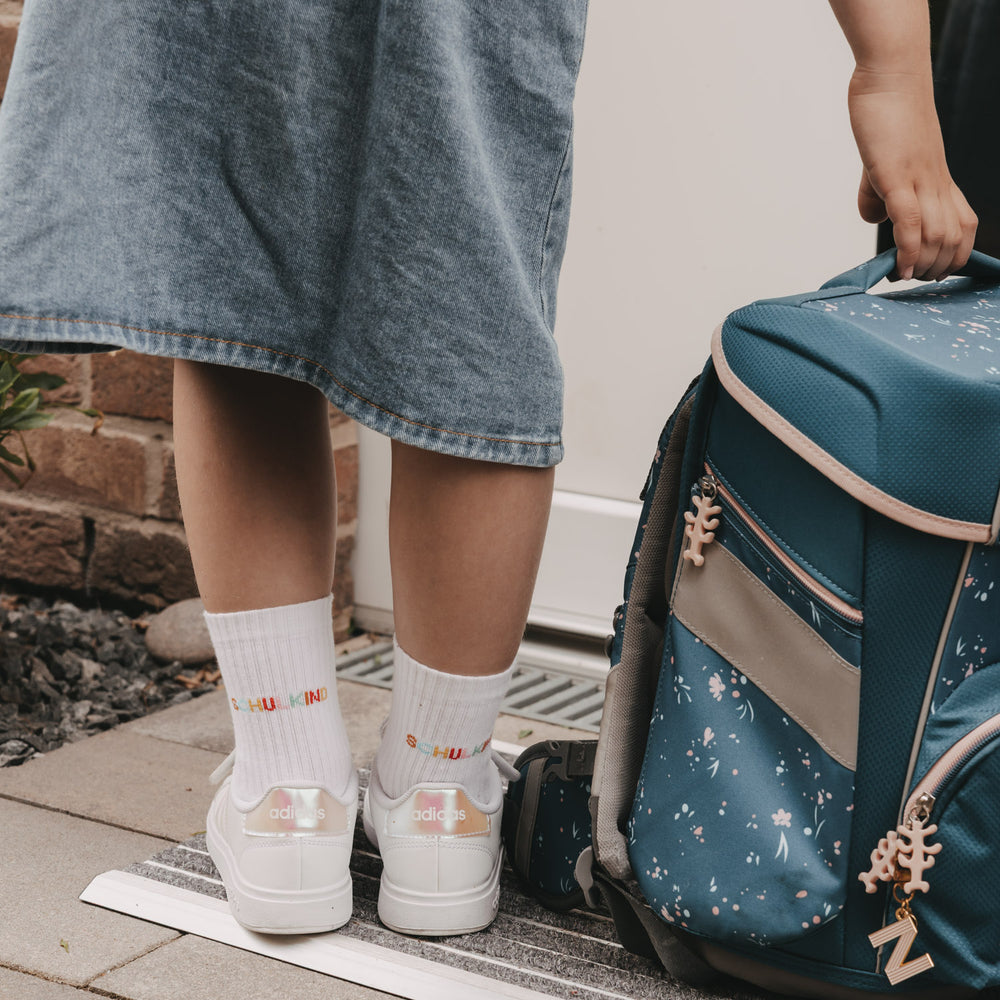 Kind mit Schulranzen trägt weiße Socken „Schulkind“ und Sneaker vor der Haustür