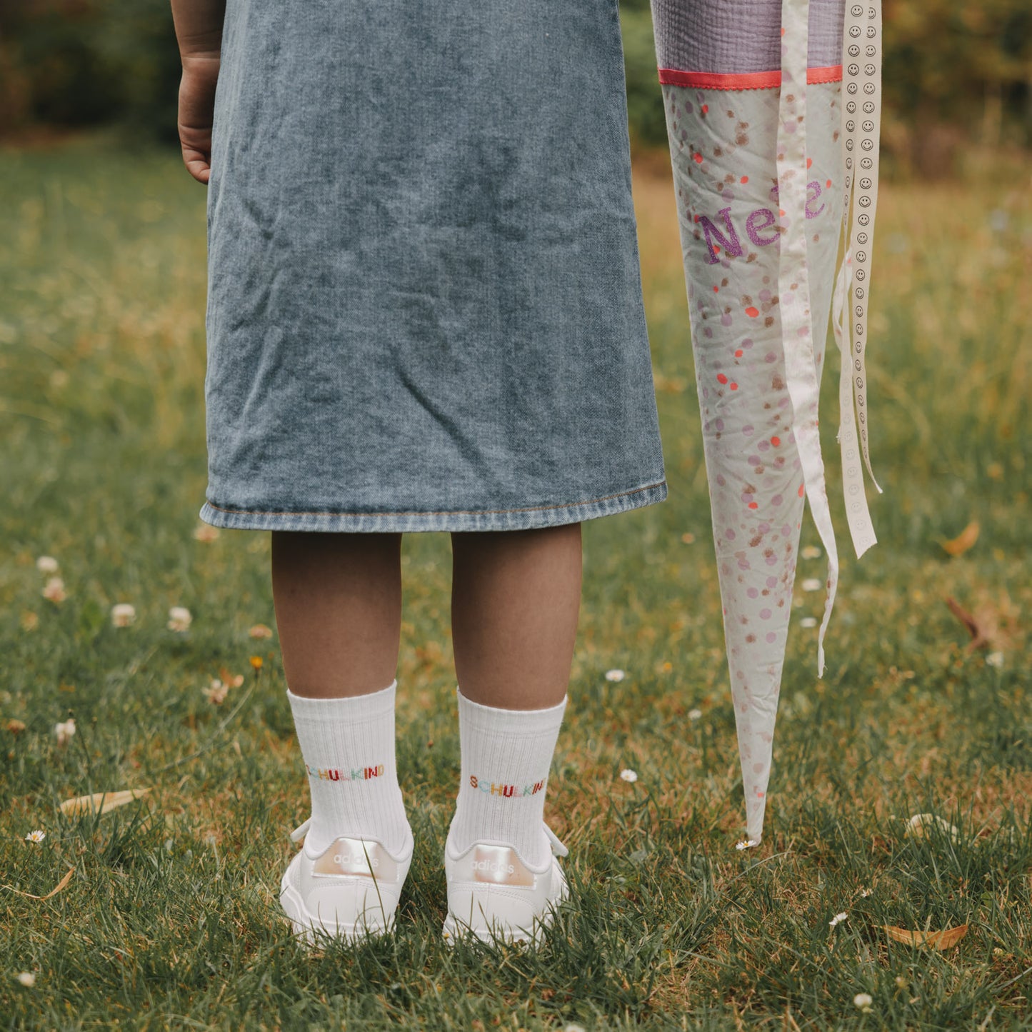 Kind mit weißen Socken „Schulkind“ und Sneakern steht auf Wiese, Schultüte daneben