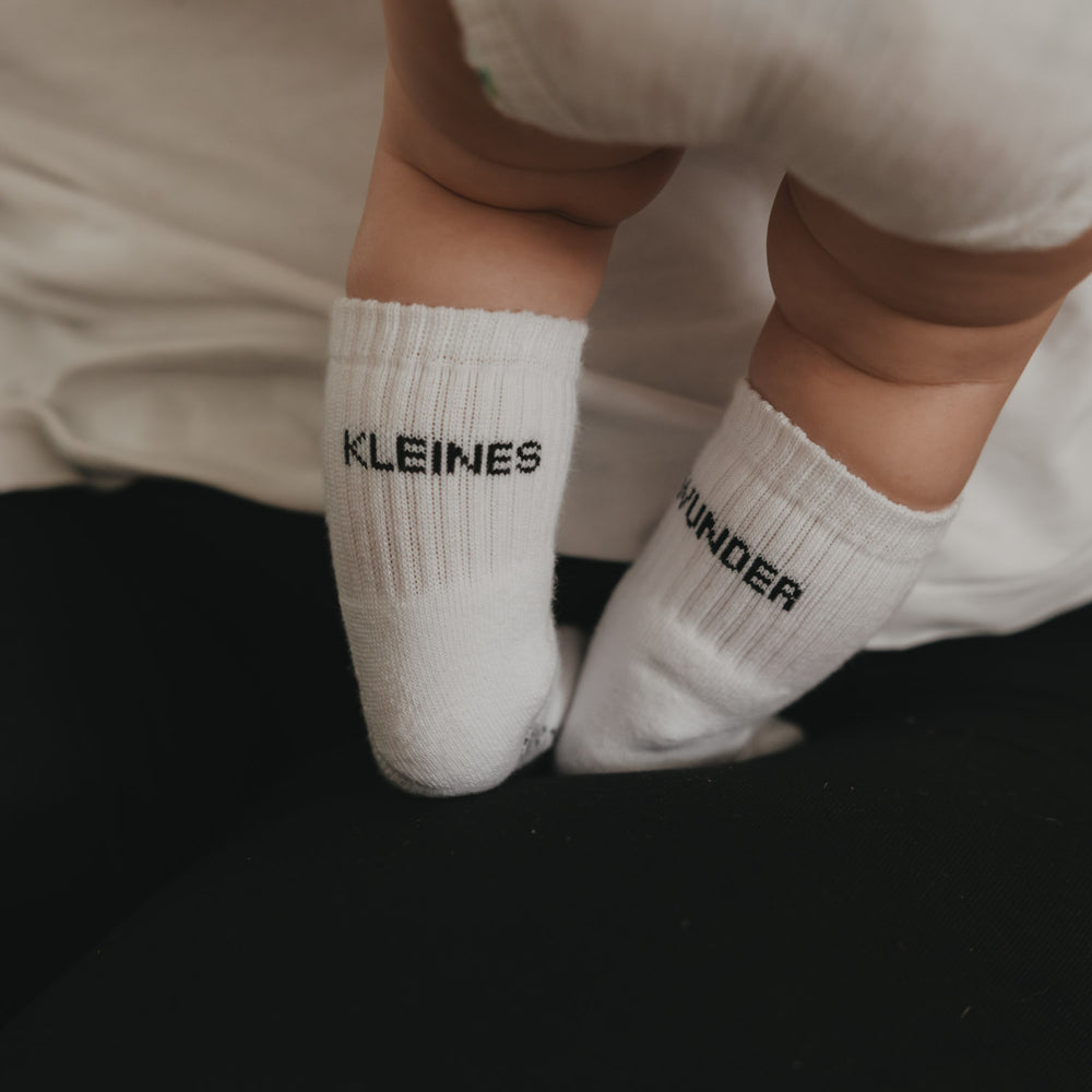 Baby trägt weiße Socken mit Schriftzug „Kleines Wunder“, Füße auf dunklem Untergrund