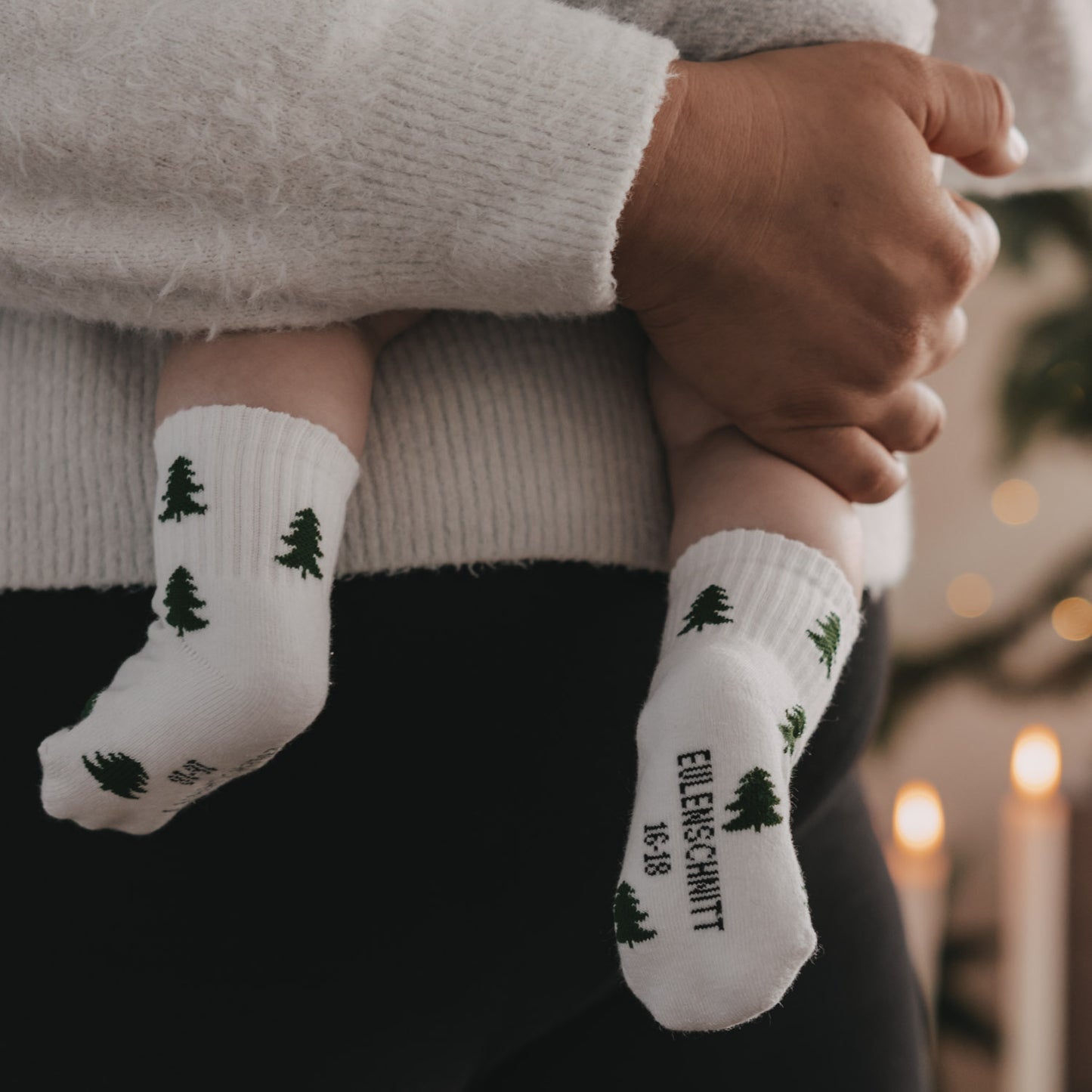 Baby trägt weiße Socken mit Tannenmotiv und Eulenschnitt Schriftzug, wird auf Arm gehalten