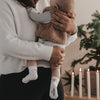 Mutter hält Baby mit weißen Socken und Schleifenmotiv, Kerzen und Tanne im Hintergrund