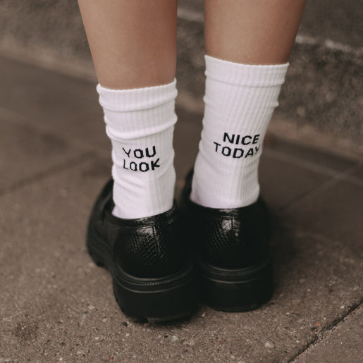 Rückansicht von Füßen in weißen Socken mit dem schwarzen Schriftzug „YOU LOOK“ und „NICE TODAY“, kombiniert mit schwarzen Loafern auf einem Gehweg.