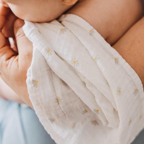 Baby wird in den Armen einer Person gehalten und in ein Musselintuch mit zartem Gänseblümchen-Muster gehüllt. Die lockere Webstruktur sorgt für eine luftige und natürliche Optik.