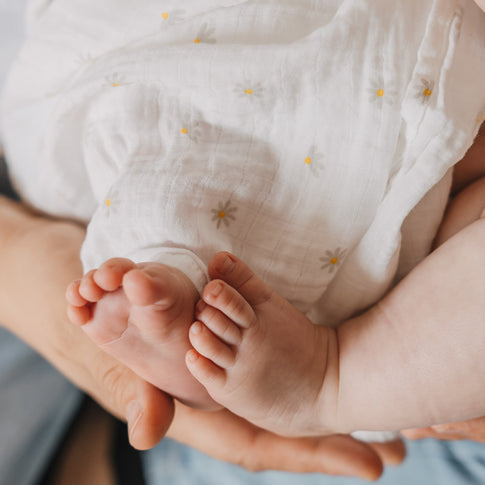 Babyfüßchen ragen aus einem weißen Musselintuch mit zartem Blumenmuster hervor. Die weiche Musselin-Qualität unterstreicht die gemütliche und liebevolle Atmosphäre.