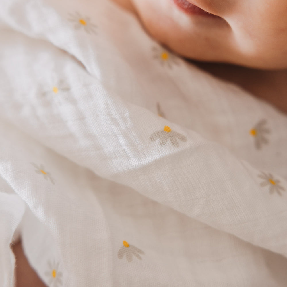 Detailaufnahme eines Babys, das in ein leichtes Musselintuch mit kleinen Gänseblümchen eingewickelt ist. Das dezente Muster verleiht dem Stoff einen verspielten und zeitlosen Charakter.
