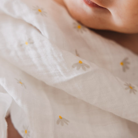 Detailaufnahme eines Babys, das in ein leichtes Musselintuch mit kleinen Gänseblümchen eingewickelt ist. Das dezente Muster verleiht dem Stoff einen verspielten und zeitlosen Charakter.