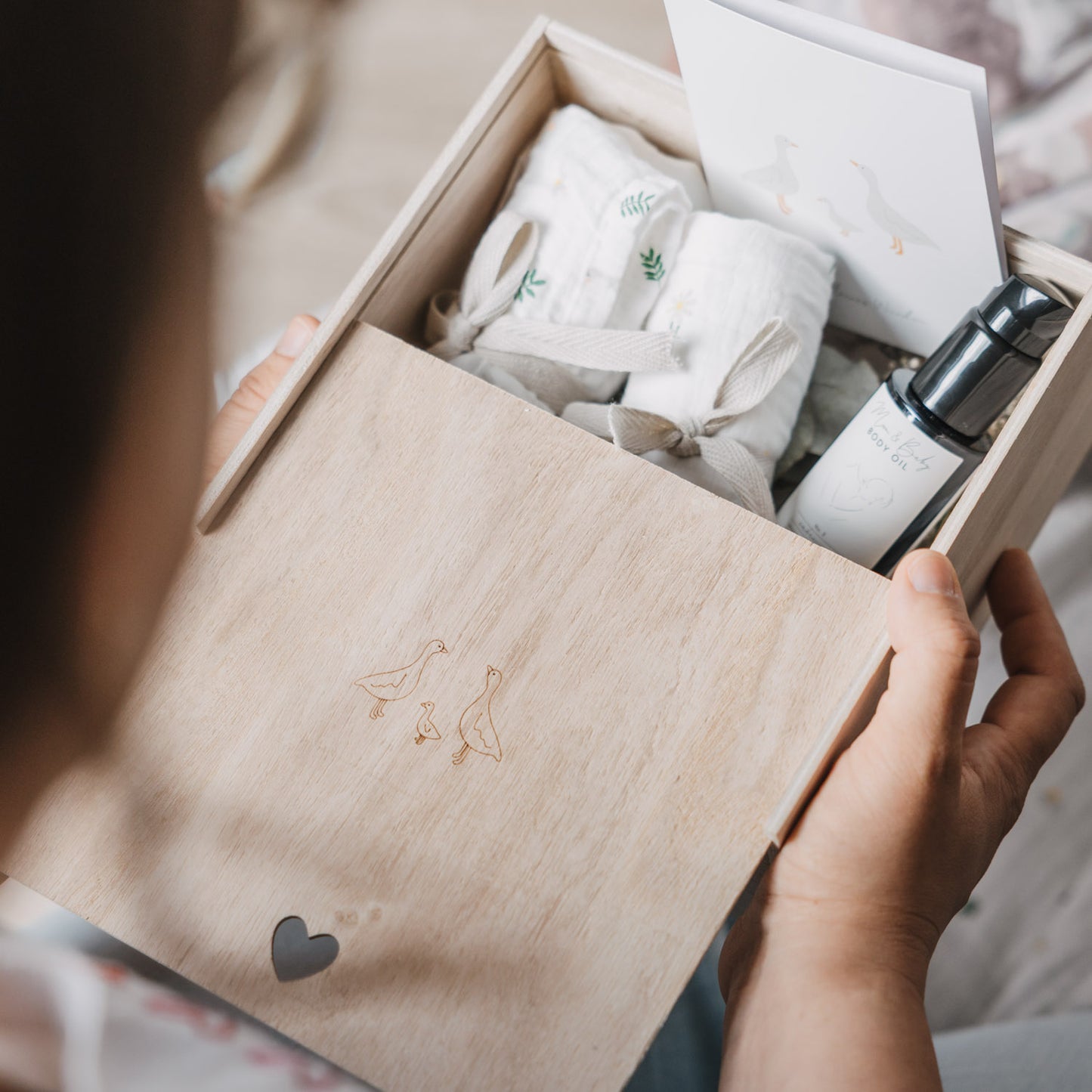 Person hält eine geöffnete Erinnerungsbox aus hellem Holz mit eingraviertem Gänsemotiv in den Händen. Im Inneren befinden sich Musselintücher mit Gänse-Print, eine Karte und ein Pflegeprodukt für Babys.