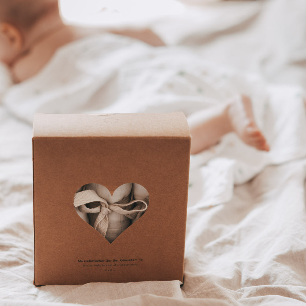 Verpackung des Musselintücher-Sets mit herzförmigem Sichtfenster auf einem Bett. Im Hintergrund liegt ein Baby eingekuschelt in den weichen Tüchern mit Gänse-Motiv.