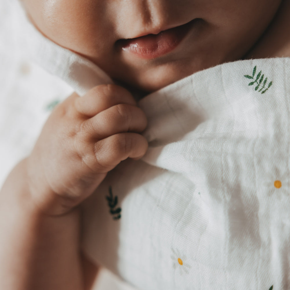 Detailaufnahme eines Babys, das ein Musselintuch mit zartem Gänse-, Blumen- und Blattmuster in der Hand hält. Die weiche Struktur des Stoffes vermittelt Geborgenheit und Komfort.