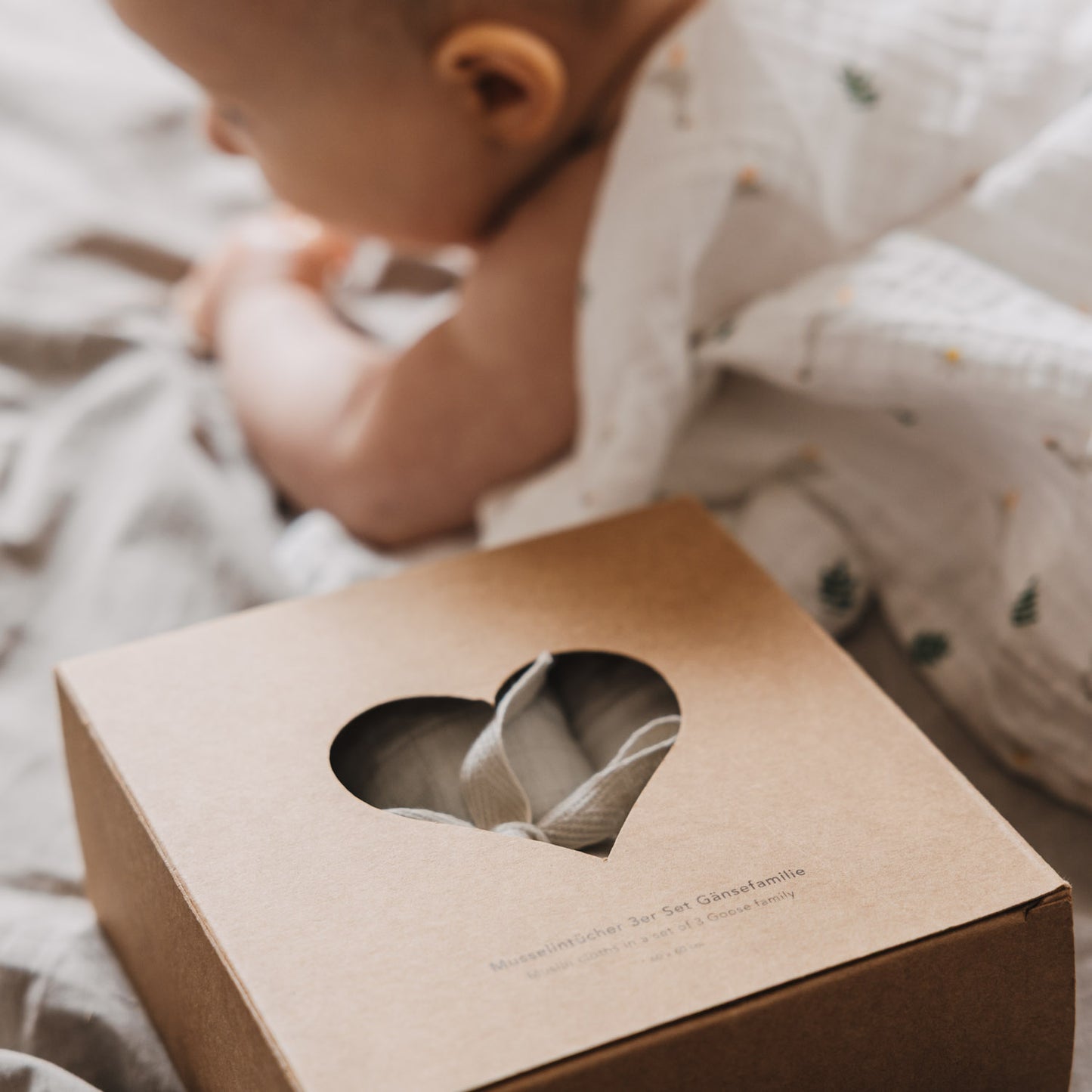 Verpackung des Musselintücher-Sets mit Herzfenster im Vordergrund, während ein Baby im Hintergrund auf einer weichen Decke liegt. Die Szene vermittelt Wärme und Geborgenheit im Babyalltag.