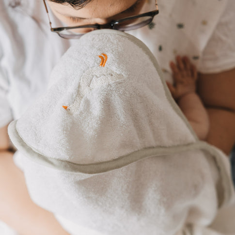 Person mit Brille hält ein Baby nach dem Baden in einem weißen Kapuzenhandtuch im Arm. Auf der Kapuze ist eine kleine gestickte Gans zu sehen, die dem Handtuch eine liebevolle Note verleiht.