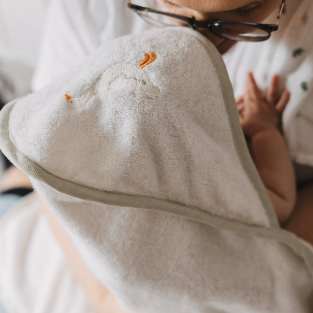 Nahaufnahme eines Babys im Arm einer Person, eingewickelt in ein weiches Kapuzenhandtuch aus Frottee. Die Kapuze mit gesticktem Gänsemotiv schützt den Kopf und sorgt für wohlige Wärme.
