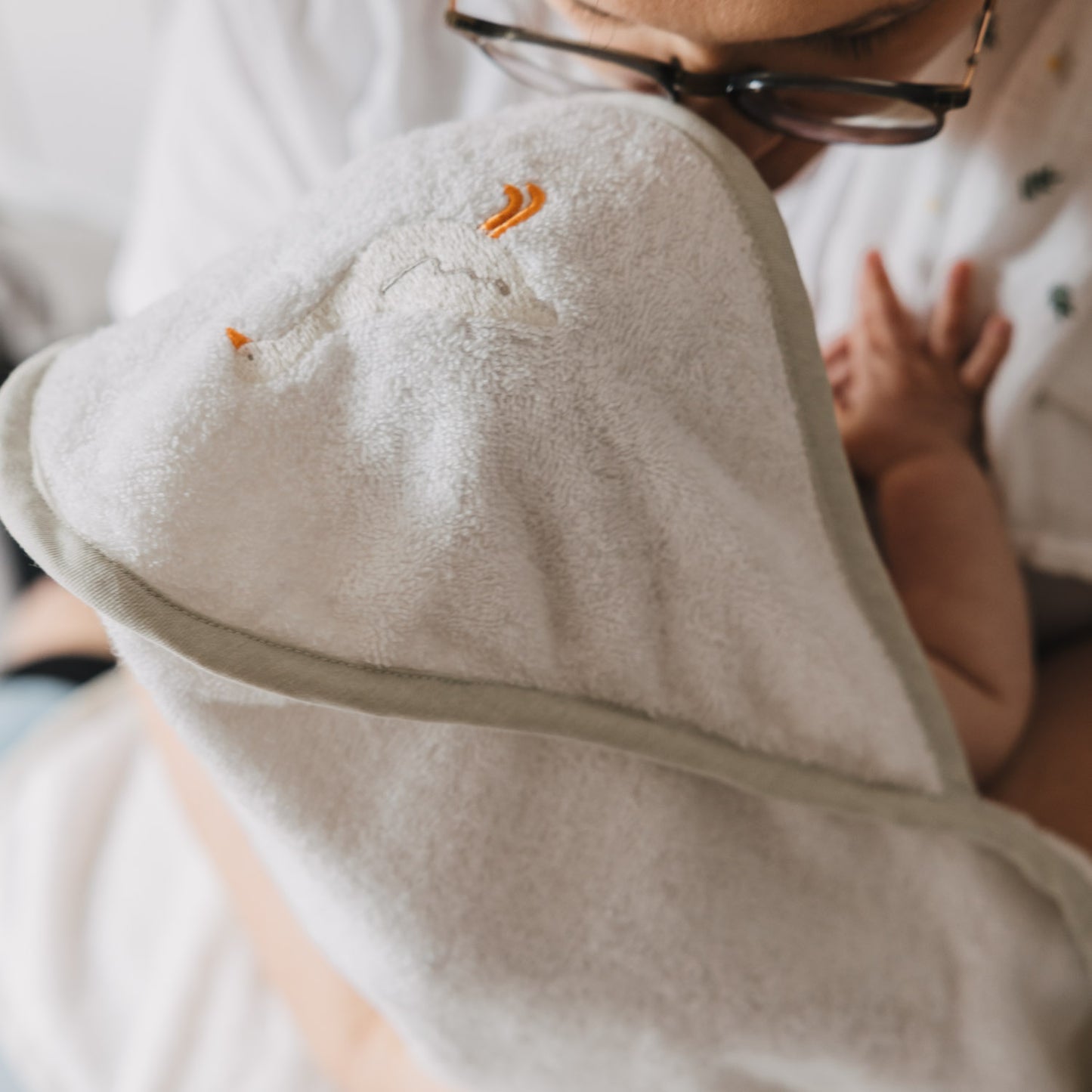 Nahaufnahme eines Babys im Arm einer Person, eingewickelt in ein weiches Kapuzenhandtuch aus Frottee. Die Kapuze mit gesticktem Gänsemotiv schützt den Kopf und sorgt für wohlige Wärme.