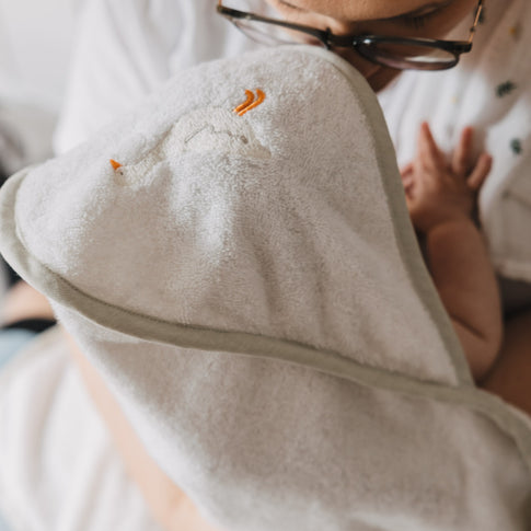 Nahaufnahme eines Babys im Arm einer Person, eingewickelt in ein weiches Kapuzenhandtuch aus Frottee. Die Kapuze mit gesticktem Gänsemotiv schützt den Kopf und sorgt für wohlige Wärme.