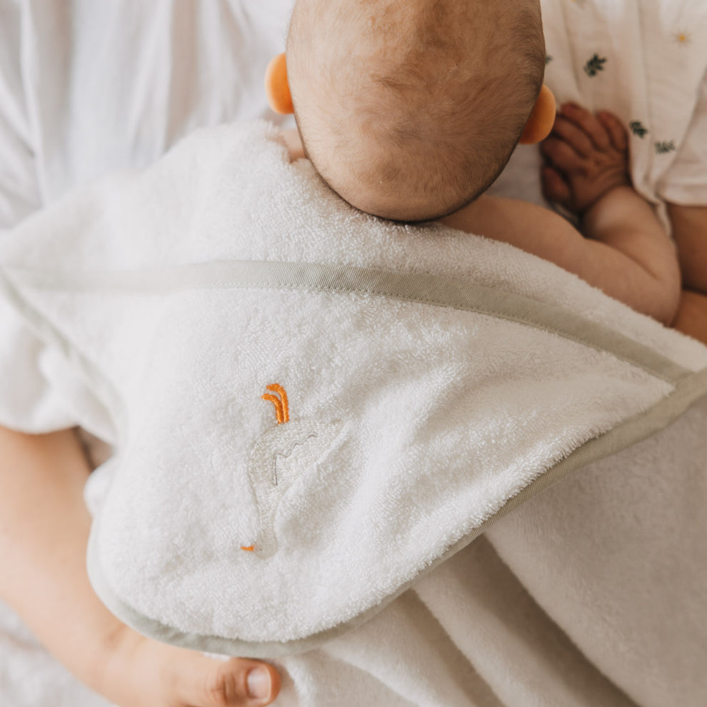 Rückansicht eines Babys, das in ein weißes Kapuzenhandtuch mit zartem Gänse-Stickmotiv eingehüllt ist. Die weiche Frottee-Qualität und die großzügige Kapuze machen das Handtuch zum idealen Begleiter nach dem Baden.