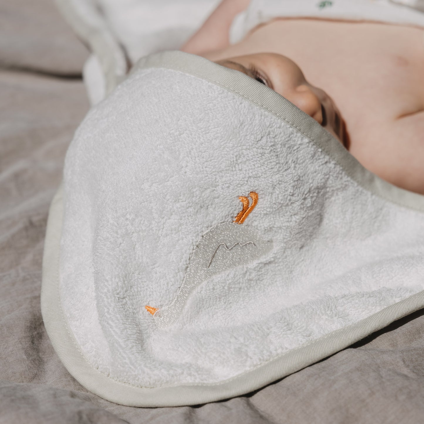 Baby liegt eingekuschelt auf einer weichen Unterlage und ist teilweise in ein weißes Kapuzenhandtuch mit dezentem Gänse-Stickmotiv gehüllt. Das flauschige Material sorgt für eine gemütliche und geborgene Atmosphäre.