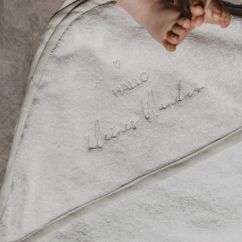 Detailaufnahme eines weißen Kapuzenhandtuchs aus weichem Frottee mit der Stickerei „Hallo kleines Wunder“ und einem kleinen Herz. Im oberen Bildbereich sind die Füßchen eines Babys zu sehen, die die liebevolle und persönliche Gestaltung des Handtuchs unterstreichen.