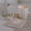 Beige Streichholzschachtel Daisy mit Blumenmotiv neben Kerzenhalter und Tasse – harmonisch inszeniert.