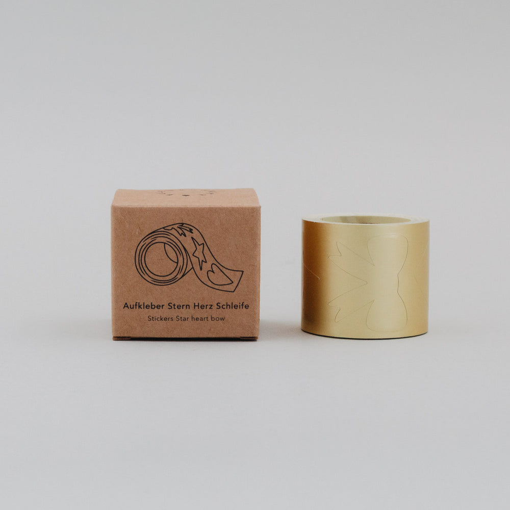 Goldene Stickerrolle mit Stern-, Herz- und Schleifenmotiv neben Verpackung auf hellem Untergrund