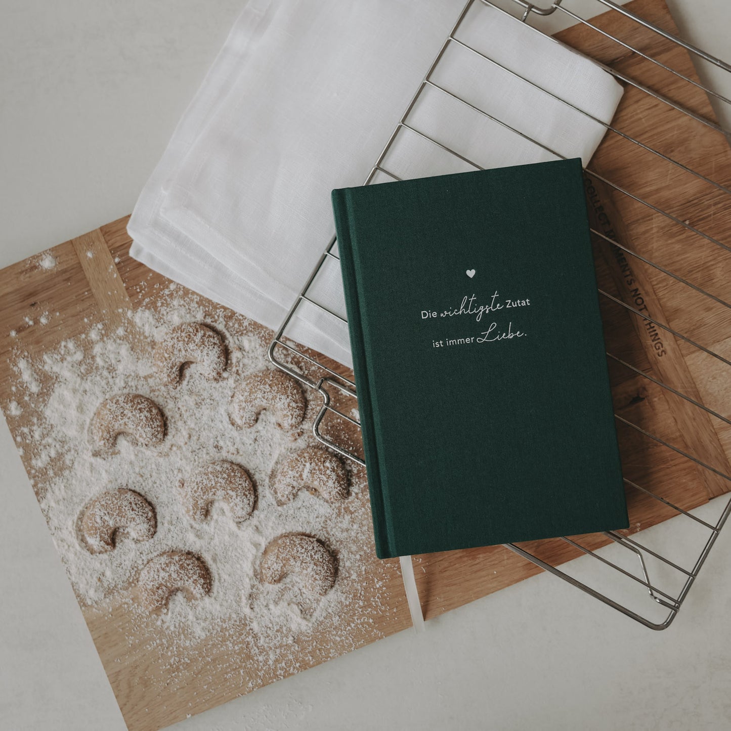 Rezeptbuch „Die wichtigste Zutat ist Liebe“ auf Backbrett mit Vanillekipferl und Kuchengitter.