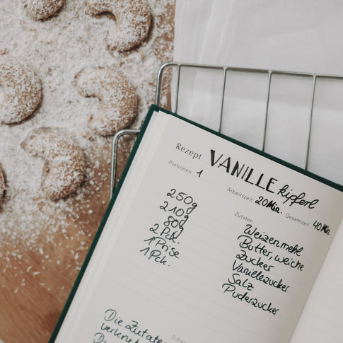 Offenes Rezeptbuch mit handgeschriebenem Vanillekipferl-Rezept neben frisch gebackenen Kipferln.