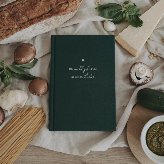 Rezeptbuch „Wichtigste Zutat“ in dunkelgrünem Einband zwischen Zutaten dekoriert