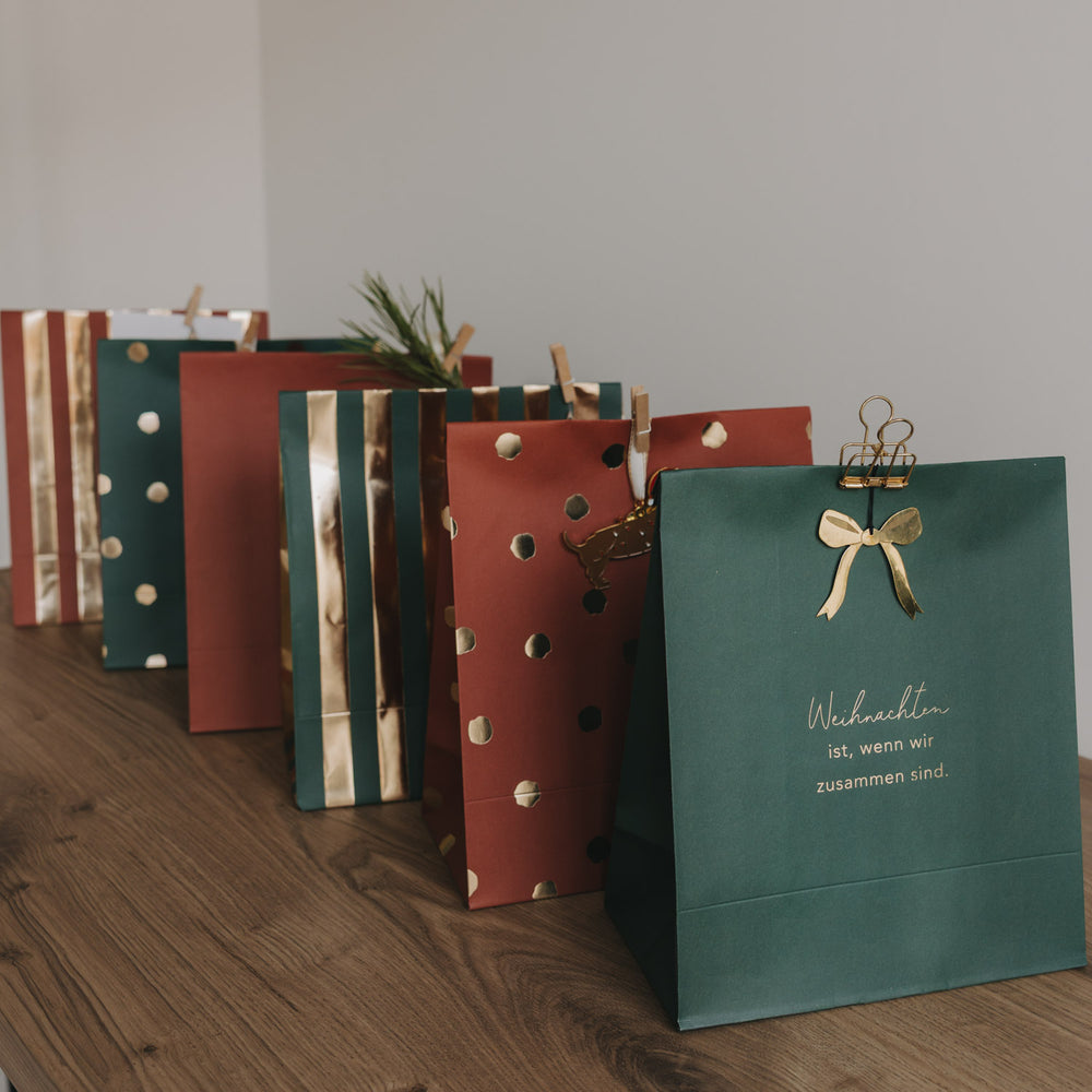 Weihnachtliche Papiergeschenktüten in Grün und Rostrot mit Golddetails, stehend arrangiert auf Holzoberfläche