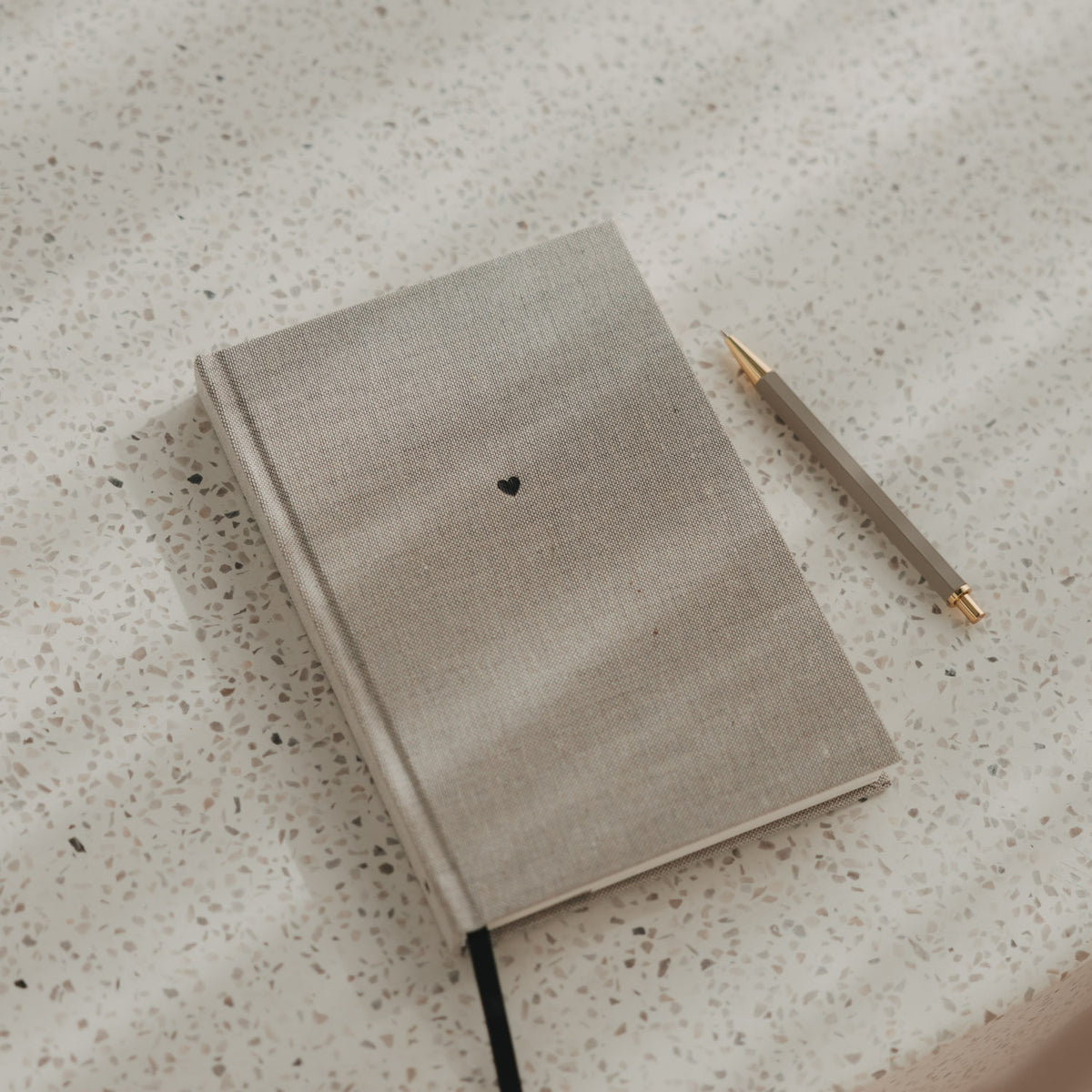 Kariertes A5 Notizbuch mit grauen Stoffeinband, schwarzem Herz und schwarzem Leseband neben einem Stift