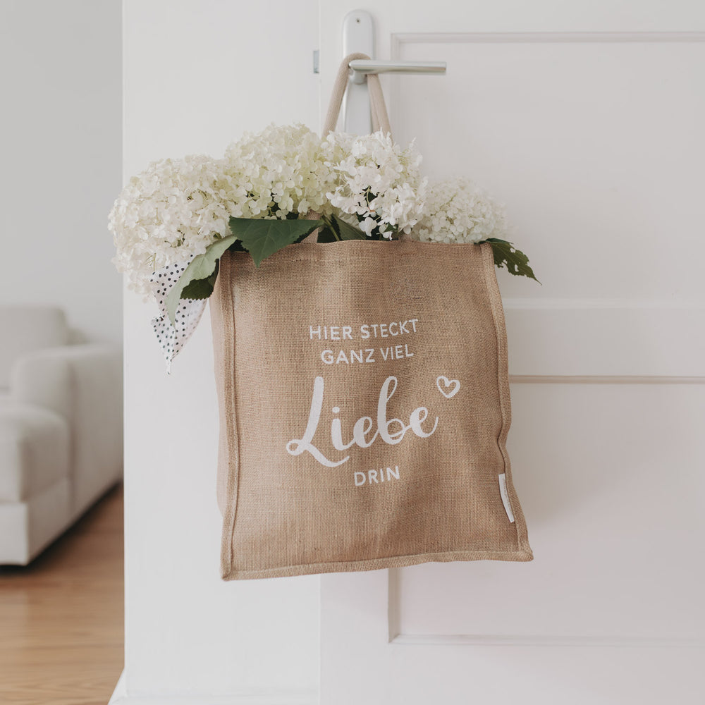 Jutetasche mit weißem Schriftzug „Hier steckt ganz viel Liebe drin“, gefüllt mit weißen Hortensien und gepunktetem Tuch, hängt an einer weißen Tür – liebevoll dekoriert.