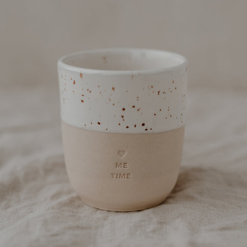 Zweifarbiger Keramikbecher „Me Time“ mit kleinem Herz und gesprenkelter Glasur auf hellem Leinenstoff.