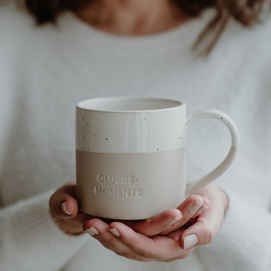 Zwei Hände halten eine beige-weiße Keramiktasse mit gesprenkelter Glasur und Henkel. Auf der Vorderseite der Tasse ist das Wort ‚GLÜCKSMOMENTE‘ eingeprägt. Die Person im Hintergrund trägt einen weichen, weißen Pullover – das Bild vermittelt Wärme, Achtsamkeit und persönliche Wohlfühlmomente.